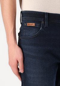 Sötétkék farmernadrág bőrszíjjal ellátott márkajelzéssel, normál zsebekkel és derékban övvel ellátott, testhezálló fazonnal.