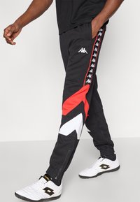 Μαύρο αθλητικό παντελόνι με κόκκινες και λευκές γεωμετρικές λεπτομέρειες, διαθέτει λογότυπο στο πλάι και φερμουάρ στα μπατζάκια για ρυθμιζόμενη εφαρμογή.