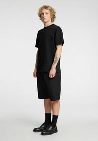 Svart bomullst-skjorte med korte ermer, kombinert med svarte knelange shorts og svarte sko, båret av en person med krøllete hår.