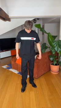 Jeune homme en pantalon noir et T-shirt noir avec une bande blanche se tient dans un salon avec un canapé orange et une grande plante en pot.