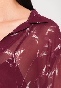 Burgundy läpikuultav blouse kaelusega, millel on valged lehemustrid. Kangas on sile ja voolav disain.