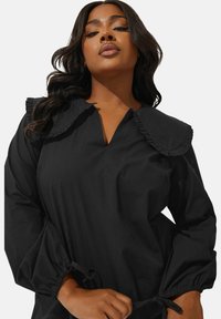 Svart klänning med volangkrage, långa ärmar med elastiska manschetter och en avslappnad passform. Tillverkad av ett slätt, lätt tyg.