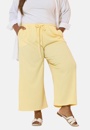 Kobieta ubrana w luźne, jasnopół żółte spodnie ze sznurkiem, białe sandały, biały top i podwiniętą białą długą koszulę, stojąca z rękami w kieszeniach.