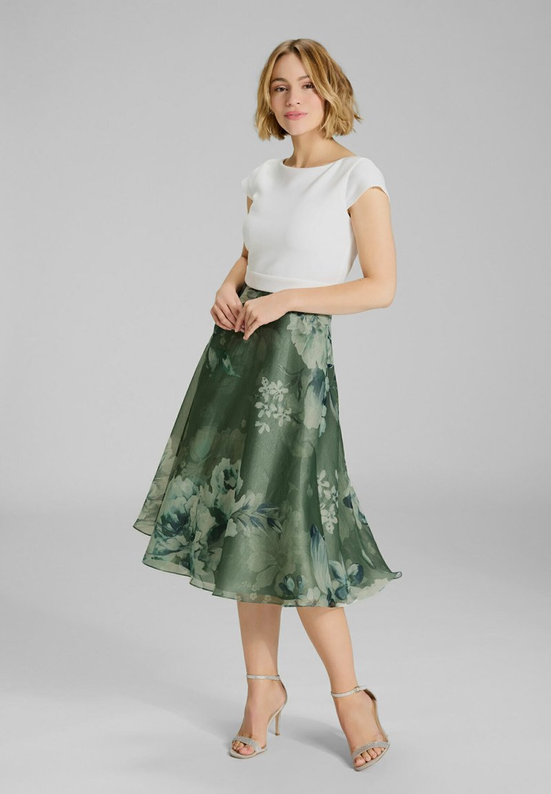 Top bianco aderente abbinato a una lunga gonna fluente verde a fiori con grandi fiori blu e bianchi. Tacchi alti con cinturini argentati completano il look.