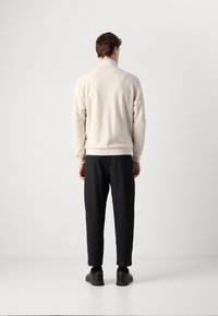 Uomo in piedi di spalle, che indossa una giacca di pile crema, pantaloni neri a cinque ottavi, calzini neri e sneakers nere contro uno sfondo bianco uniforme.