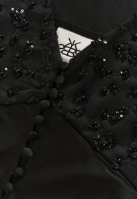 Crna odjeća s teksturiranom tkaninom ukrašenom šljokicama i dekorativnim gumbima, ističući složene detalje i glatku podstavu.