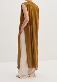 Bezrekawnikowa, długa brązowa narzutka o zwiewnym kroju. Lekki, półprzezroczysty materiał, boczne rozcięcia i luźny krój na kremowych spodniach.