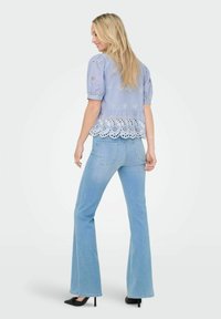 Blouse bleu clair avec des motifs brodés et un ourlet ondulé, associée à un jean bleu clair évasé et des chaussures à talons noirs.