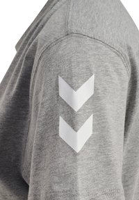 Grå t-shirt med rund hals. Venstre ærme har to hvide chevronformer. Materialet er blødt og tekstureret med synlige sømme.