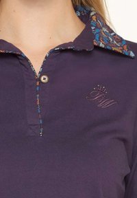 Polo shirt violet foncé avec un col à motifs floraux. Il présente une fermeture à boutons sur le devant et un logo brodé sur la poitrine. Texture douce et lisse.