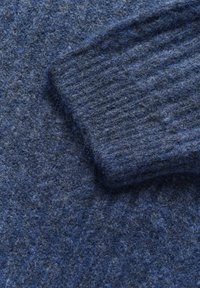 Maglione blu in lana con lavorazione a maglia spessa e polsini a costine, che presenta un aspetto morbido e peloso con tonalità di blu variabili.
