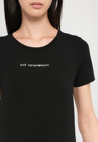 Svart t-skjorte med korte ermer laget av glatt stoff, med en hvit logo "EA7 EMPORIO ARMANI" trykket på brystet.