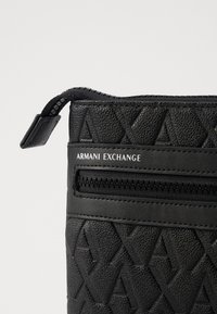 Ova crna teksturirana faux kožna torbica s geometrijskim utisnutim uzorkom ima zatvarač i brendirana je s "Armani Exchange."