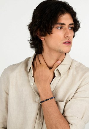 Bracciale di perline con perline blu e blu navy, caratterizzato da un mix di design lisci e testurizzati. Indossato su un polso abbronzato con una camicia beige.