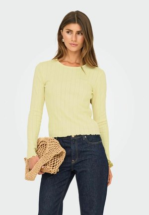 Pastelgul ribbad långärmad topp med rundad halsringning och vågig fåll, tillsammans med mörk denimjeans och en strukturerad beige väska.
