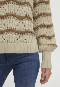 Strickpullover in Creme mit braunen Streifen, präsentiert mit strukturierten Rautenmuster und gerippten Bündchen. Getragen mit blauen Denim-Jeans.