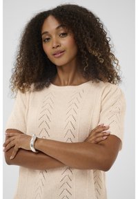 Ženska s kodrastimi lasmi, oblečena v svetlo roza pleteno majico s kratkimi rokavi, roke prekrižane, ter srebrna zapestnica na njenem zapestju pred enobarvno ozadje.