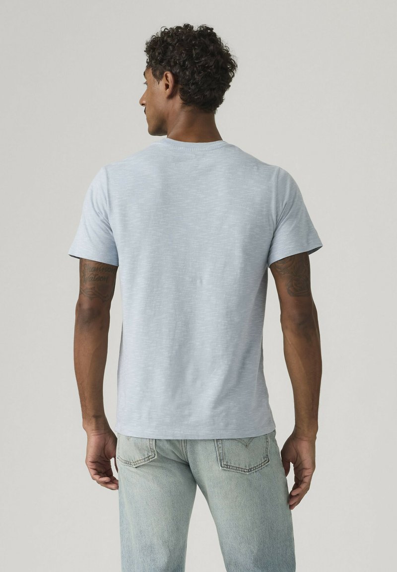 Uomo con capelli ricci che indossa una maglietta a maniche corte azzurro chiaro e jeans azzurri, in piedi di spalle alla fotocamera su uno sfondo semplice.