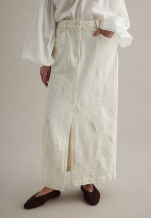 Donna che indossa una lunga gonna crema con uno spacco frontale, abbinata a una camicetta bianca con maniche a sbuffo e scarpe slip-on marrone scuro.