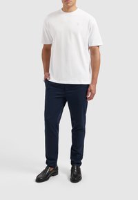 Witte t-shirt met korte mouwen met een subtiel logo, gecombineerd met marineblauwe broek en zwarte instappers, wat een casual maar verzorgde uitstraling geeft.