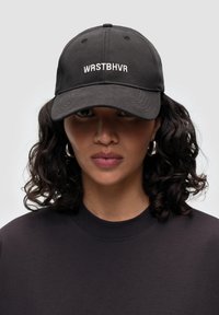 Svart basebollkeps med broderad "WRSTBHV" i vitt, i bomullsmaterial, med rundad krona och böjd skärm. Bärs av en person med lockigt hår.