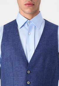 Colete texturizado azul sobre uma camisa colarinho clara azul, apresentando três botões escuros e um decote em V.