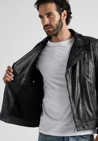 Schwarze Leder-Motorradjacke mit silbernen Reißverschlüssen, Druckknopfdetails und einer strukturierten Oberfläche, getragen über einem schlichten weißen T-Shirt.