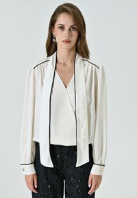 Blusa bianca con scollatura a V profonda, maniche lunghe, piping nero e colletto drappeggiato. Abbinata a pantaloni neri con paillettes e dettagli con bottoni dorati.