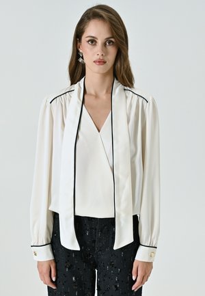 Blusa bianca con scollatura a V profonda, maniche lunghe, piping nero e colletto drappeggiato. Abbinata a pantaloni neri con paillettes e dettagli con bottoni dorati.