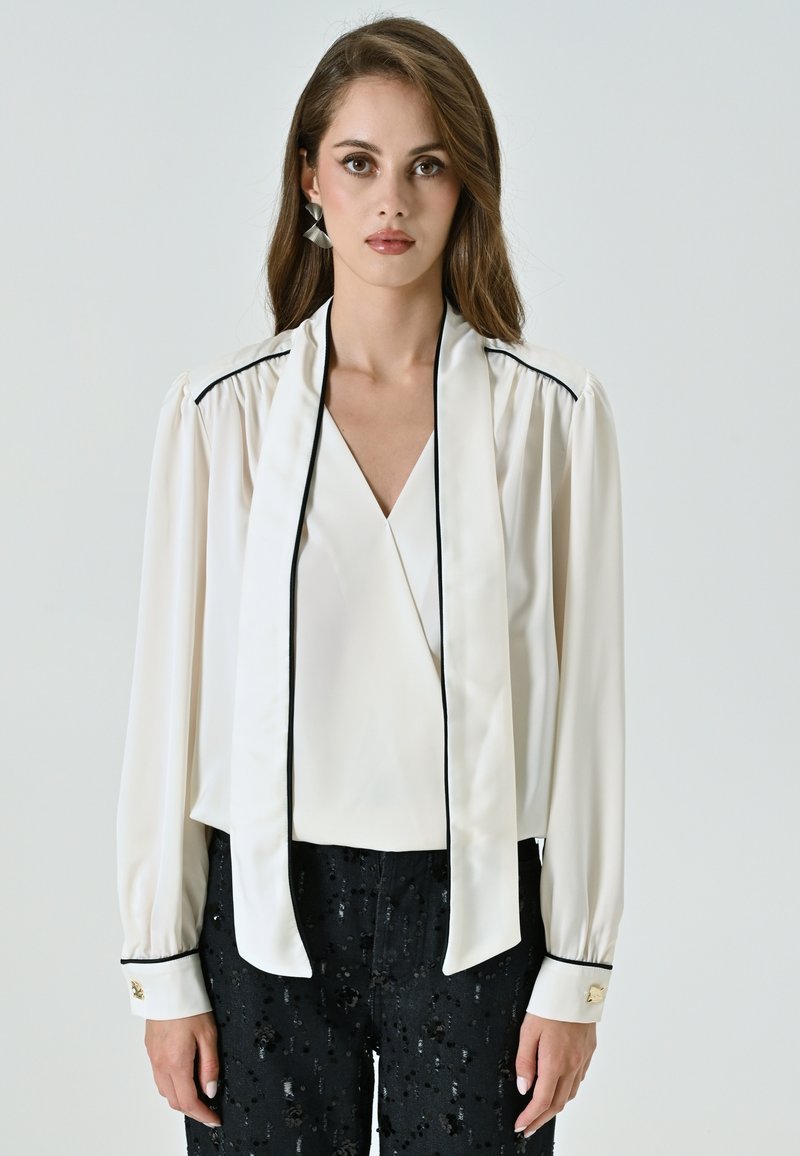 Blusa bianca con scollatura a V profonda, maniche lunghe, piping nero e colletto drappeggiato. Abbinata a pantaloni neri con paillettes e dettagli con bottoni dorati.