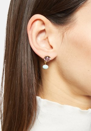 Goudkleurige oorbellen met een ronde paarse steen en een druppelvormige aqua-accent, hangend aan een stud, weergegeven aan het oor van een model.