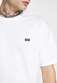 Camiseta blanca de algodón con cuello redondo y mangas cortas. Presenta un pequeño parche negro del logo de Vans en el área del pecho izquierdo.