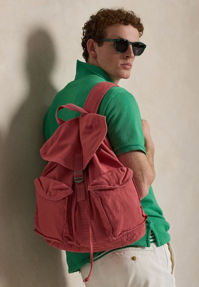 Άνδρας που φοράει σκουρόχρωμα γυαλιά ηλίου, πράσινο πόλο μπλουζάκι, λευκό παντελόνι και κουβαλάει ένα μεγάλο κόκκινο καμβά σακίδιο, στέκεται μπροστά σε μια μπεζ τοίχο.