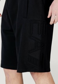 Pantaloni corti sportivi neri realizzati in tessuto morbido, con motivi geometrici in rilievo sui lati e vita con cordoncino.