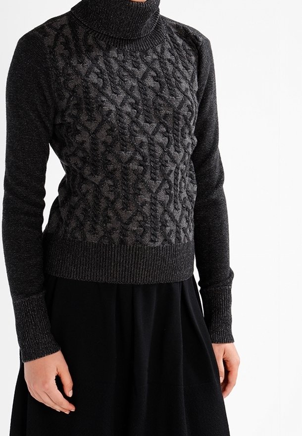 Personne portant un pull à col roulé gris foncé à torsades avec manches longues et une jupe noire, se tenant devant un fond blanc.