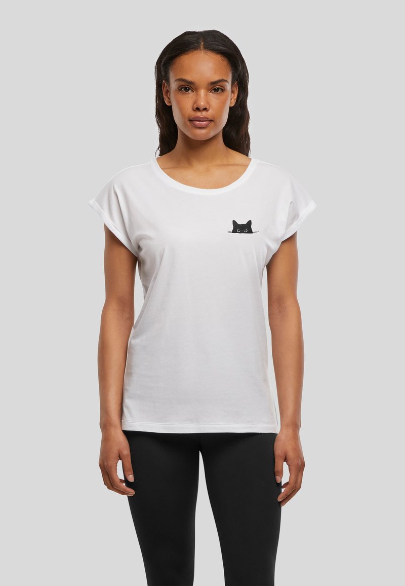 Femme portant un T-shirt blanc avec un petit motif de chat noir près de la poitrine gauche, associé à un pantalon noir, debout devant un fond uni.