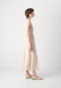 Beige sleeveless dress with a defined waist, flowing material, and ankle length. Model paired with light-colored flat shoes.