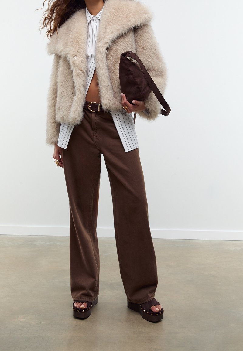 Femme portant une veste en fausse fourrure beige, une chemise rayée, un pantalon marron à jambes larges, des sandales à plateforme, tenant un sac à main marron foncé contre un mur blanc.