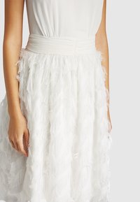 Robe blanche avec un corsage ajusté et une jupe à volants. La jupe présente une texture douce et légère comme des plumes, ainsi qu'une taille lisse.