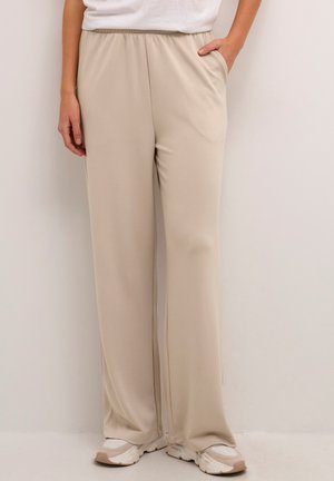 Pantalon beige à jambes larges avec ceinture élastique, deux poches latérales et texture de tissu lisse, associé à des baskets de couleur claire.