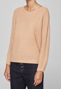 Lichtbeige gebreide trui met een boothals, lange mouwen en een losse pasvorm. De textuur is licht geribbeld, wat goed combineert met donkere spijkerbroeken.