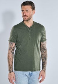 Olive green short-sleeve Henley shirt in cotton blend with five-button placket, round neck, and stitched hem. Features visible arm tattoos.