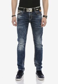 Blaue Jeans aus Denim mit schlankem Schnitt, die ausgebleichte Stellen, Kontrastnähte und seitliche Akzente aufweisen. Getragen mit einem schwarzen Gürtel und Sneakers.