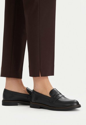 Czarne skórzane wsuwane mokasyny noszone z ciemnobrązowymi skróconymi spodniami odsłaniającymi kostkę i boczne rozcięcie.