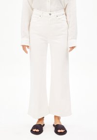 Jeans blancs à jambes larges avec un design à cinq poches, fermeture à bouton en métal et couture discrète. Portés avec des sandales noires à enfiler.