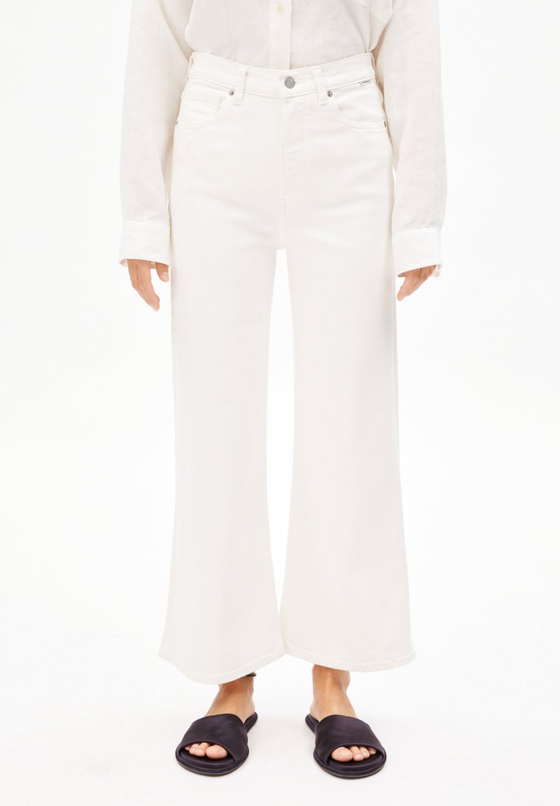 Jeans blancs à jambes larges avec un design à cinq poches, fermeture à bouton en métal et couture discrète. Portés avec des sandales noires à enfiler.