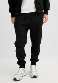 Pantalones jogger negros hechos de tela suave, con cintura ajustable con cordón y paneles laterales, combinados con zapatillas deportivas blancas.