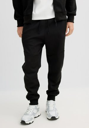Pantalones jogger negros hechos de tela suave, con cintura ajustable con cordón y paneles laterales, combinados con zapatillas deportivas blancas.