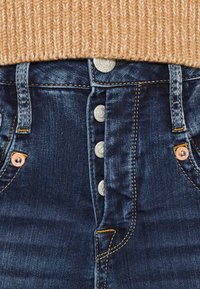 Mörkblå denimjeans med fem silverknappar, orange sömmar och kopparnitar. Texturerad midjeband under en ljusbrun stickad tröja.