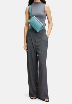 Sac en cuir teal porté en bandoulière avec une fermeture éclair, doté d'une finition texturée et d'un logo en relief, porté sur un haut gris ajusté et un pantalon large gris.